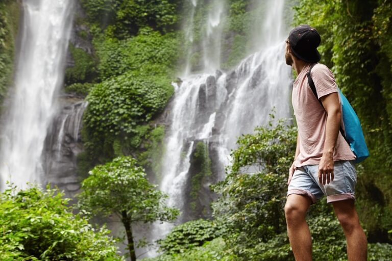 People, wildlife and adventure concept. Fashionable young adventurer with backpack contemplating waterfall landscape. Male tourist enjoying beautiful nature around him during his journey in rainforest